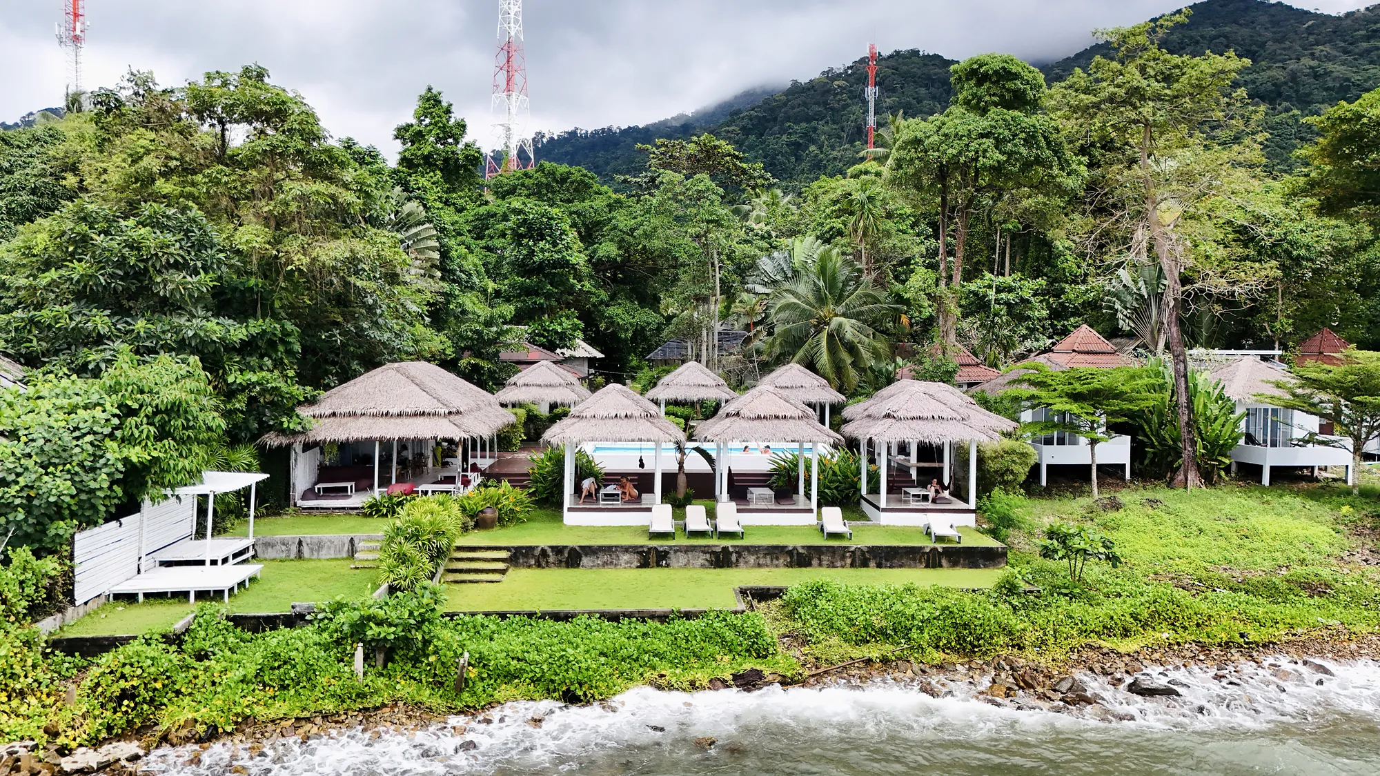 The resort from the water: waves on the rocks, the lawn and the thatched salas below the jungle